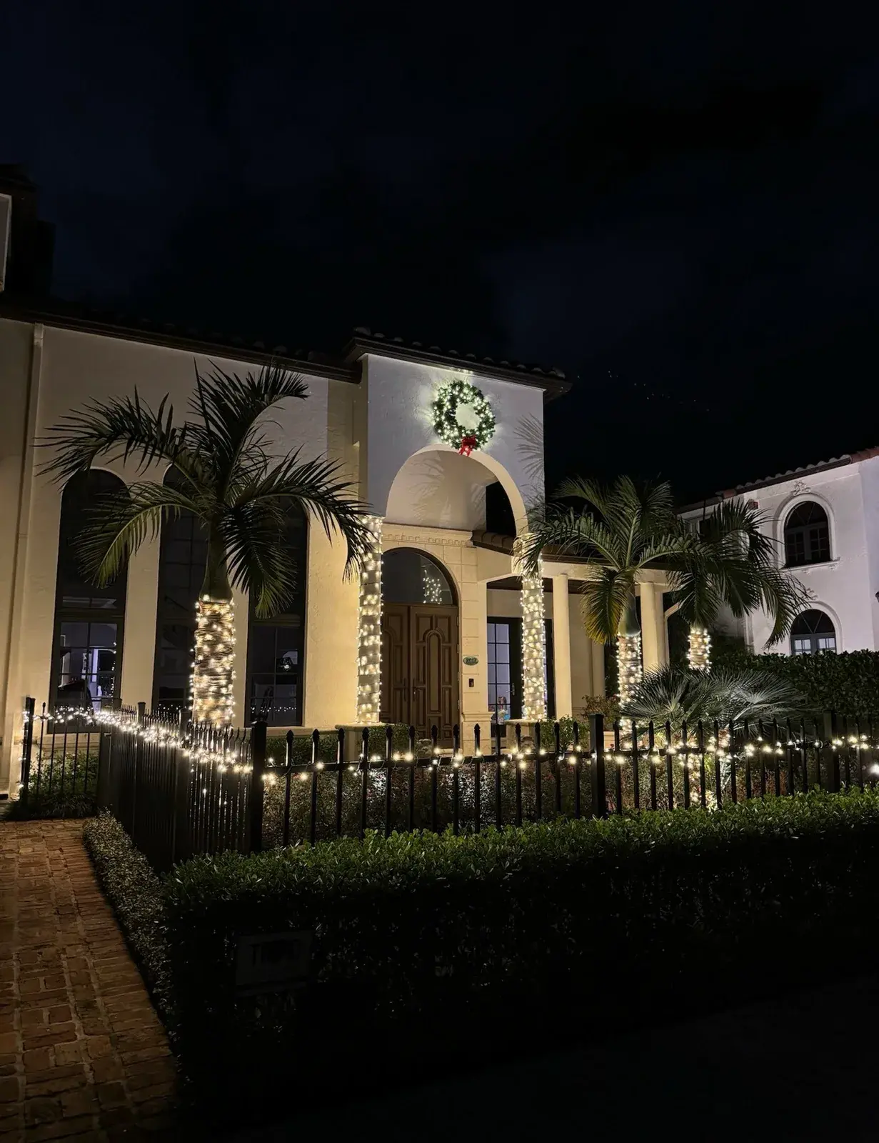 Mediterranean-style home with warm white holiday lights, palm trees, and a lit wreath