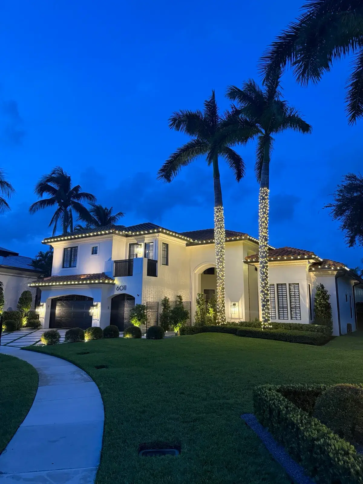 White two-story home with roofline lights and illuminated palm trunks at blue hour